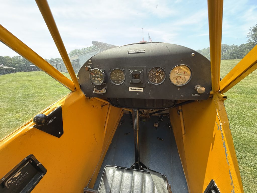 N6625H J3 Cub 1946 - Countrywide Flyers | Orlando Apopka X04 View from the pilot’s seat in a bright yellow small airplane, showing a round gauge cluster and control panel with a grassy field outside the open canopy.