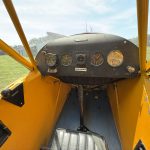 View from the pilot’s seat in a bright yellow small airplane, showing a round gauge cluster and control panel with a grassy field outside the open canopy.