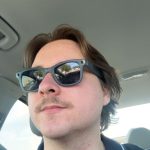 Close-up selfie of a man wearing blue sunglasses in a car, seatbelt across chest, reflection of trees/road in the lenses.