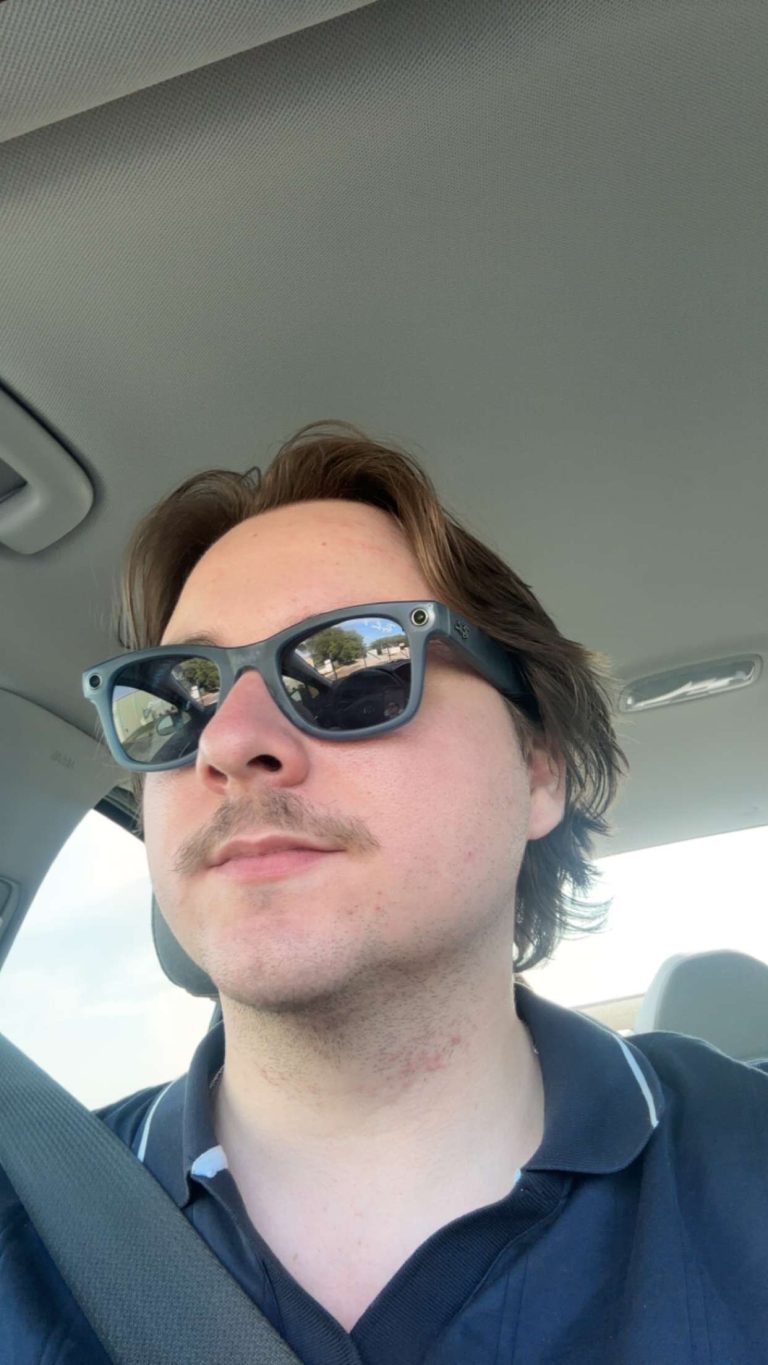 Close-up selfie of a man wearing blue sunglasses in a car, seatbelt across chest, reflection of trees/road in the lenses.