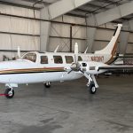 White twin-engine turboprop aircraft in a hangar, orange-gold stripe along the fuselage, red towing strap on the nose gear.
