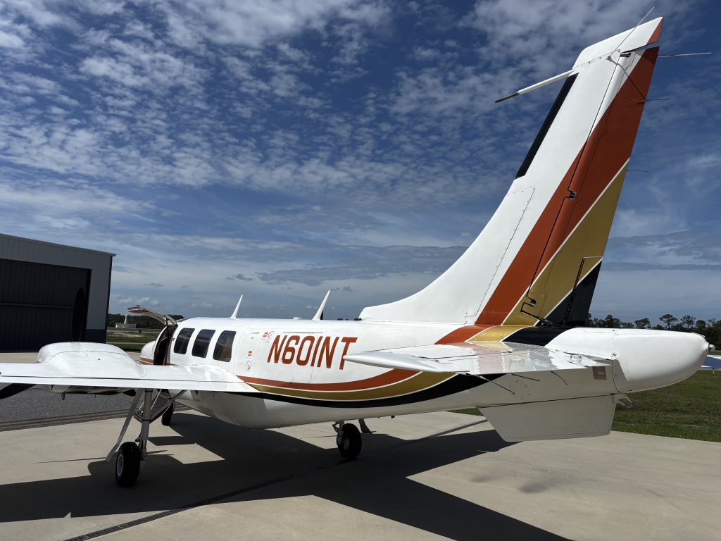 Small white airplane with orange and yellow tail parked on a tarmac, hangar on the left and a blue sky with clouds in the background.