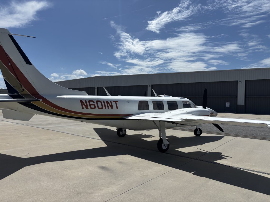 Tail and fuselage of a small white private airplane with red and gold accents on a tarmac, hangar in background under a blue sky with wispy clouds.