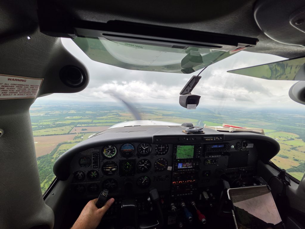 Pilot's left hand on control yoke, cockpit instrument panel visible as scenery passes outside the windshield above fields and roads.