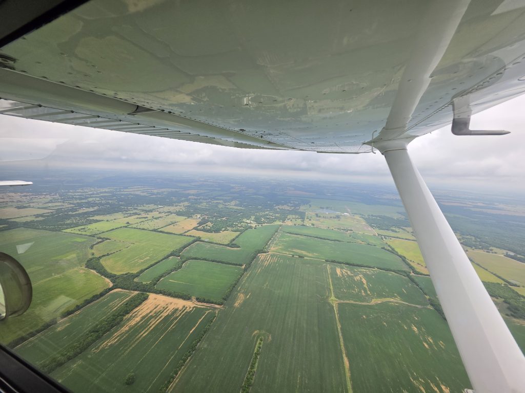 Aerial view of patchwork farmland and fields seen from a small plane window, with wing strut in frame above.