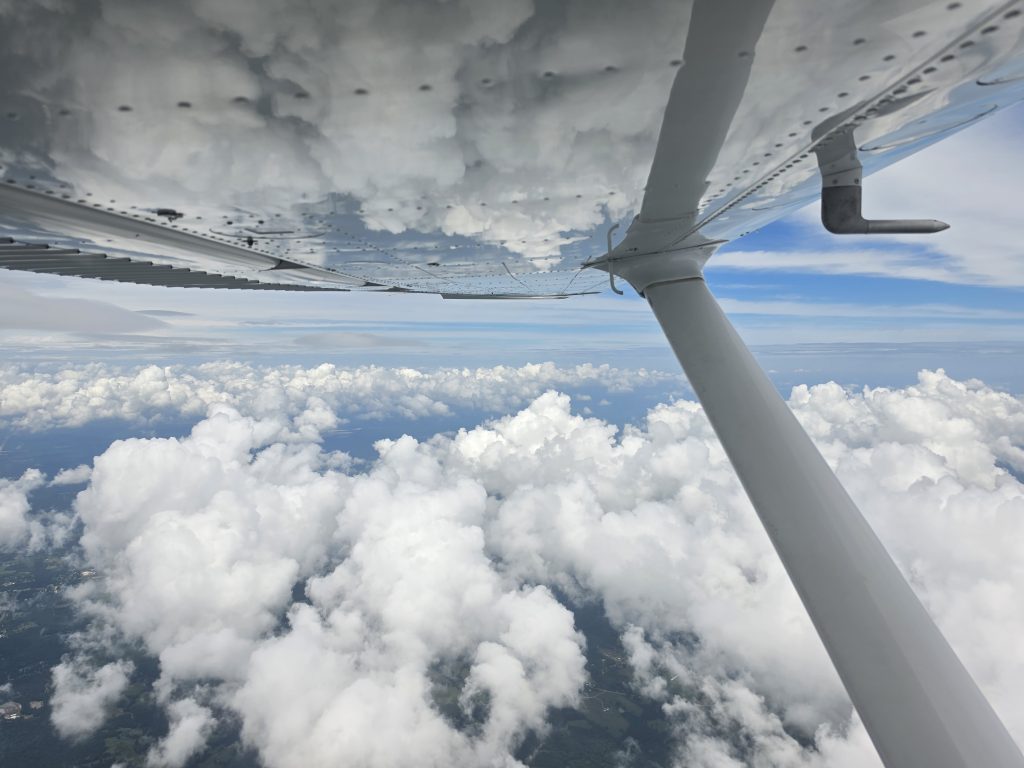 Passenger-window view of an airplane wing and strut, flying above a sea of white clouds.