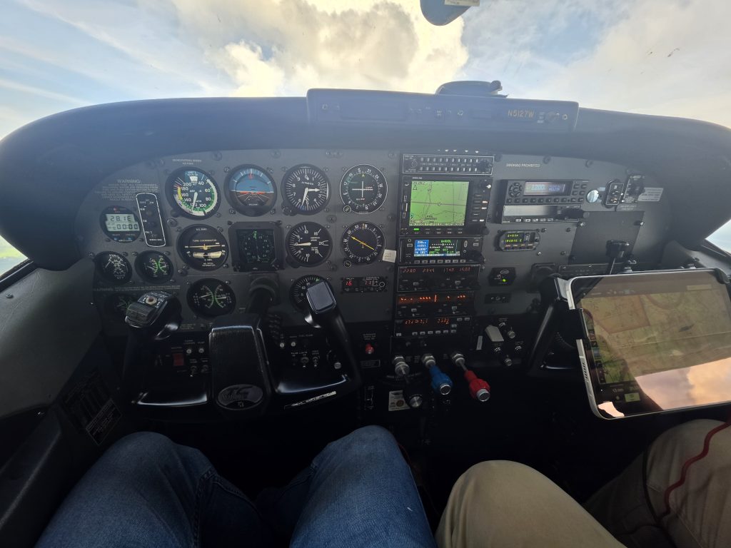 Cockpit of a small aircraft with multiple gauges and navigation screens; pilot's legs visible in jeans and a map tablet on the right side.