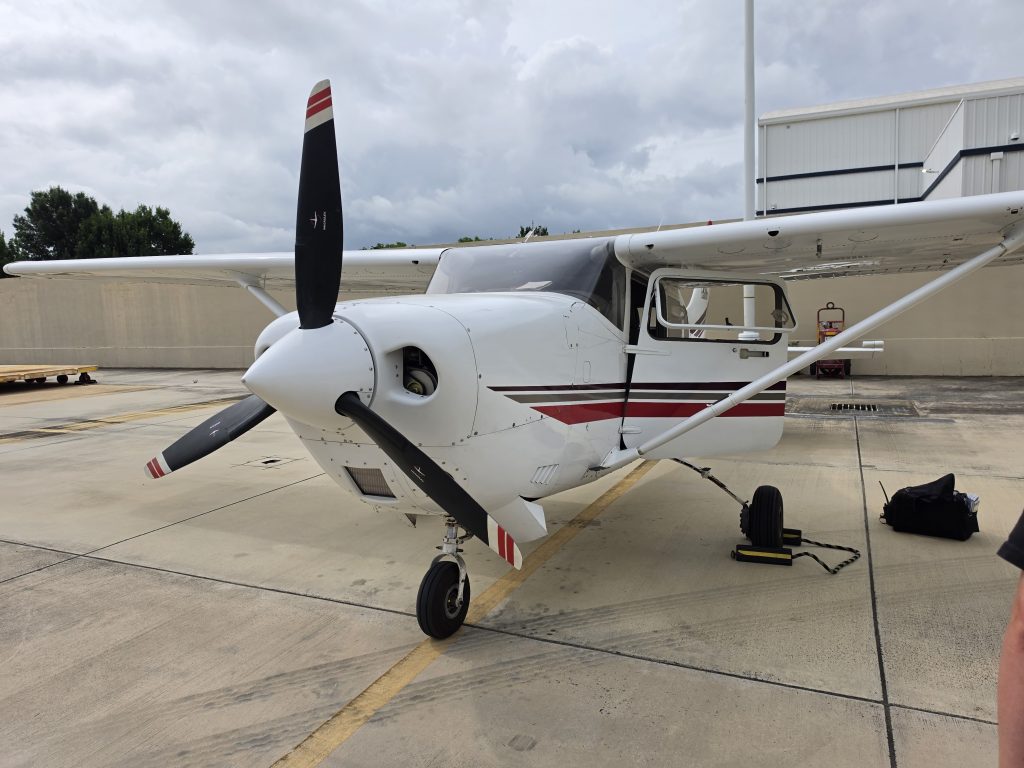 Small white single-engine airplane parked on a concrete tarmac with its propeller visible and a hangar in the background under a cloudy sky.