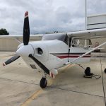 Small white single-engine airplane parked on a concrete tarmac with its propeller visible and a hangar in the background under a cloudy sky.