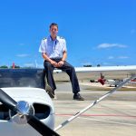 Pilot in uniform sits on the wing of a small propeller plane on tarmac, blue sky above.