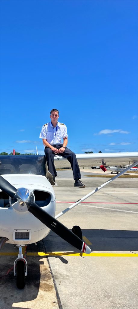 Hayes Barnett 1 - Countrywide Flyers | Orlando Apopka X04 Pilot in uniform sits on the wing of a small propeller plane on tarmac, blue sky above.