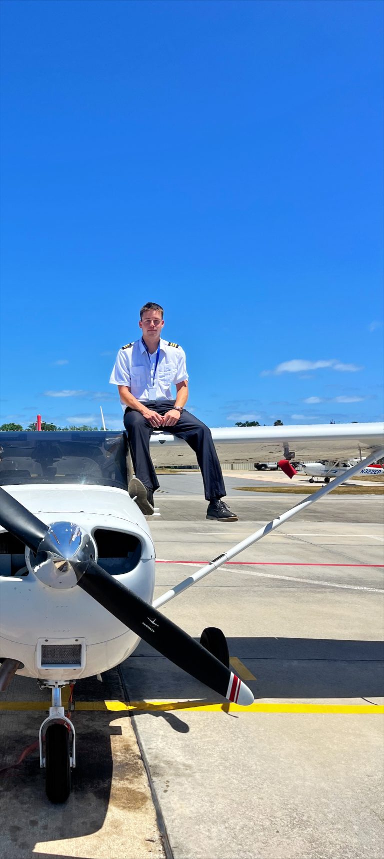 Pilot in uniform sits on the wing of a small propeller plane on tarmac, blue sky above.