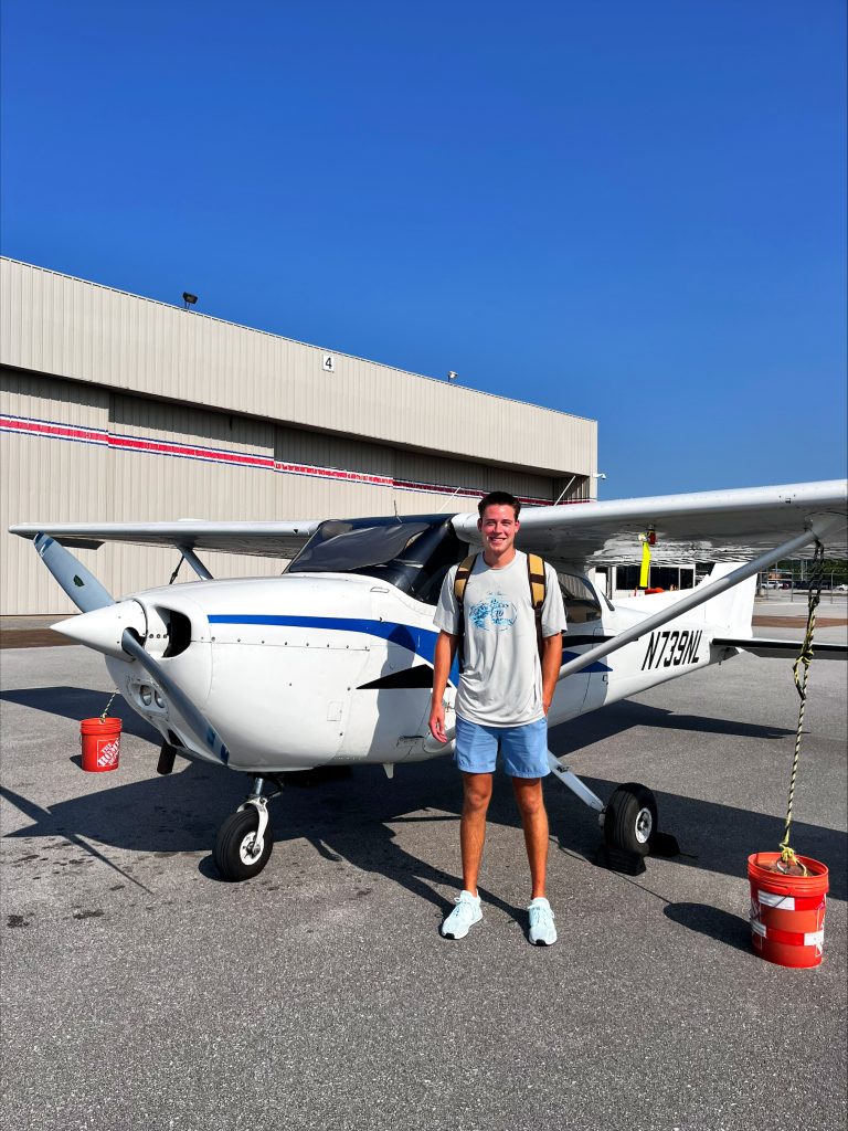 Hayes Barnett 2 - Countrywide Flyers | Orlando Apopka X04 Young man in a light t‑shirt and backpack stands beside a small white airplane on a sunny tarmac, with a hangar and bright blue sky in the background.