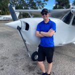 Man in blue polo shirt and cap posing with folded arms beside a small white single-engine airplane on a sunny day.