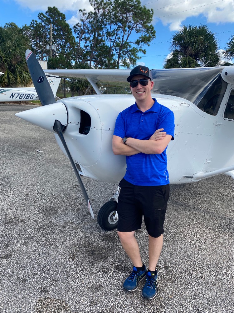 Jordan McNeel - Countrywide Flyers | Orlando Apopka X04 Man in blue polo shirt and cap posing with folded arms beside a small white single-engine airplane on a sunny day.