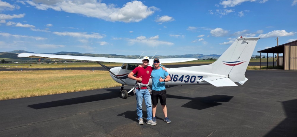 Jordan McNeel 2 - Countrywide Flyers | Orlando Apopka X04 Two people pose with a small white airplane on a runway, blue sky and hills in the background, a sunny day.