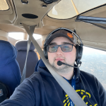 Man wearing a headset with microphone in a small airplane cockpit, seatbelt fastened, window view of the landscape outside.