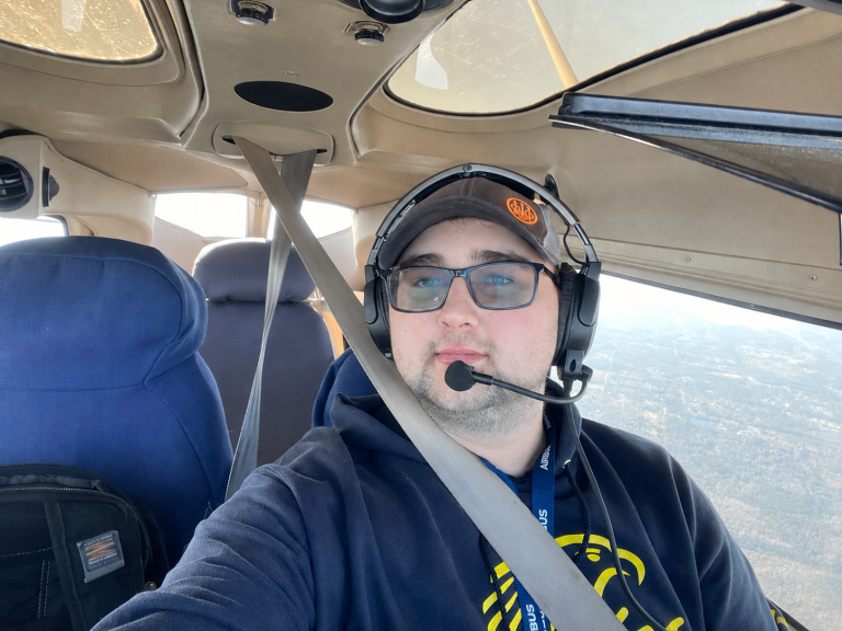 Man wearing a headset with microphone in a small airplane cockpit, seatbelt fastened, window view of the landscape outside.