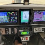 Cockpit of a small aircraft with three large digital screens showing flight instruments and navigation.