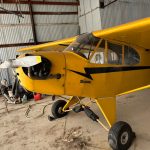 Yellow vintage single-engine airplane in a hangar with the engine exposed and propeller in front, landing gear visible.
