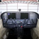 Interior of a small airplane cockpit with dual yokes, two large electronic flight displays, and central throttle controls