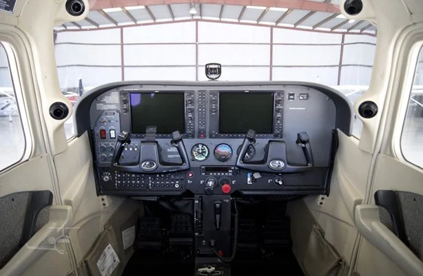 Interior of a small airplane cockpit with dual yokes, two large electronic flight displays, and central throttle controls