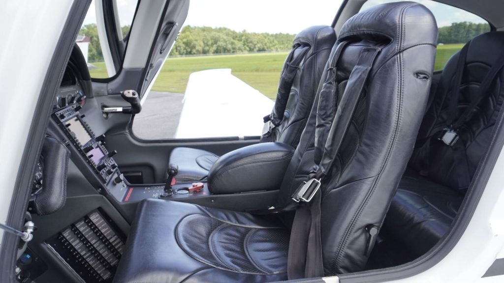 Front cockpit of a small helicopter with leather seats, joystick controls, and a view of the runway outside the open door.