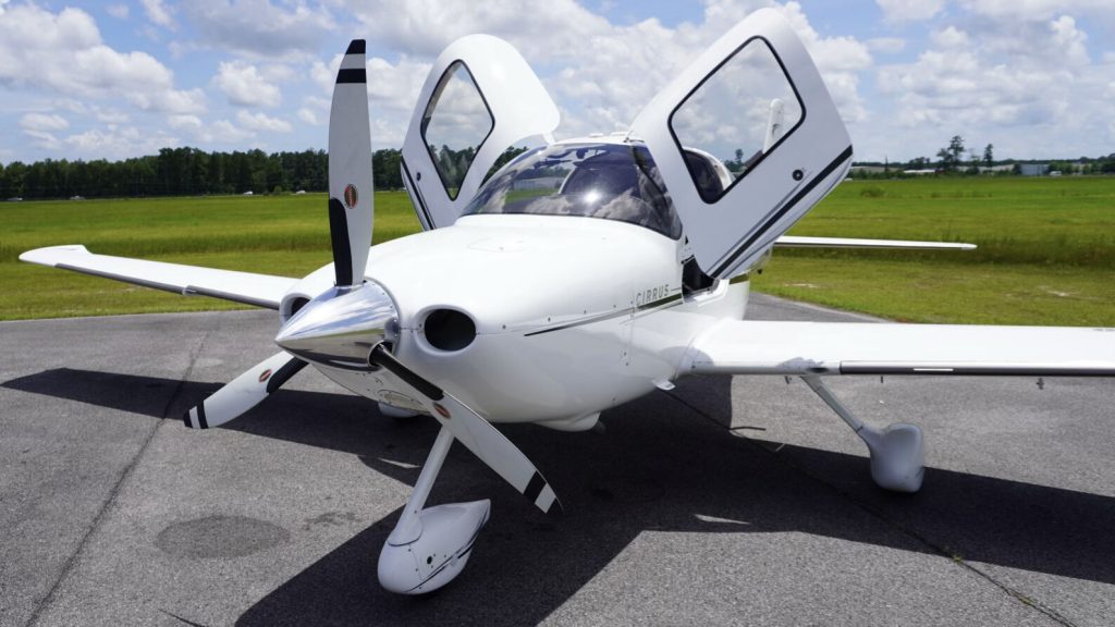 White Cirrus private airplane parked on a paved ramp with its gull-wing doors open and propeller at rest, green fields and blue sky in the background