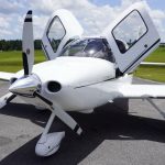 White Cirrus private airplane parked on a paved ramp with its gull-wing doors open and propeller at rest, green fields and blue sky in the background