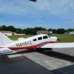 Small white and red single-engine airplane parked on a concrete apron with a grassy field and houses in the background; registration N4161Z