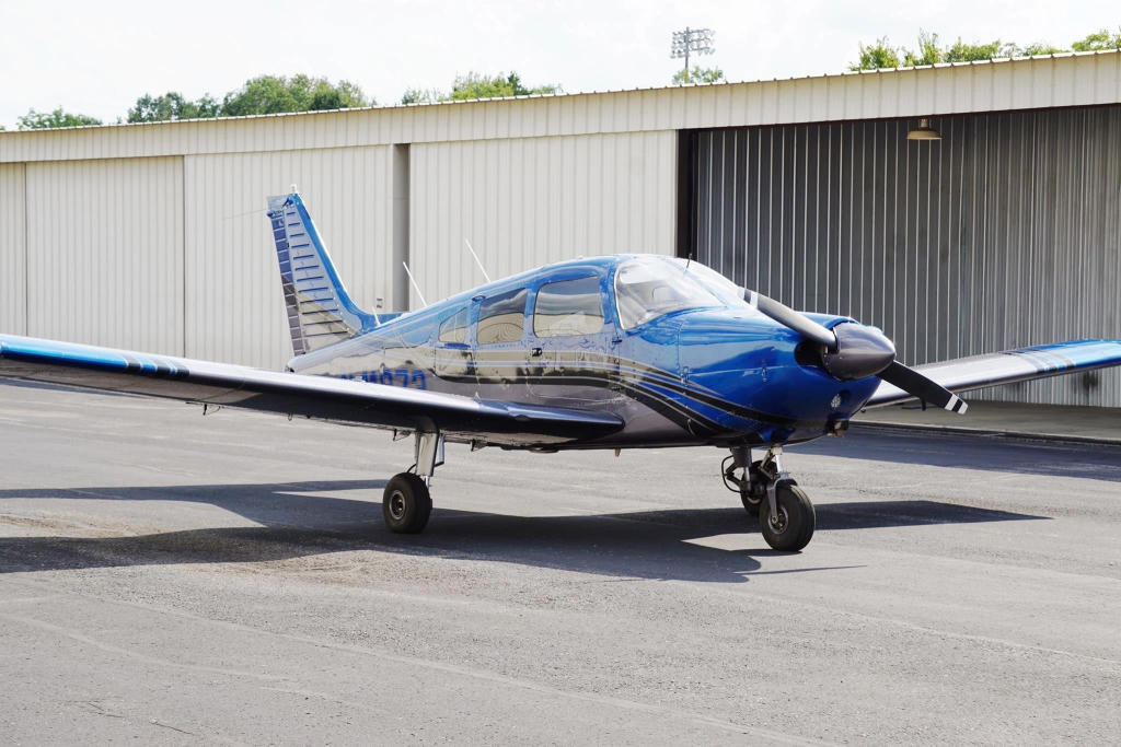 Blue single-engine propeller airplane parked on the tarmac in front of a white hangar building.