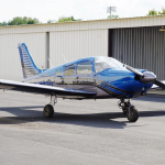 Blue single-engine propeller airplane parked on the tarmac in front of a white hangar building.