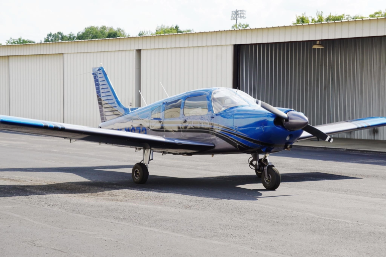 Blue single-engine propeller airplane parked on the tarmac in front of a white hangar building.