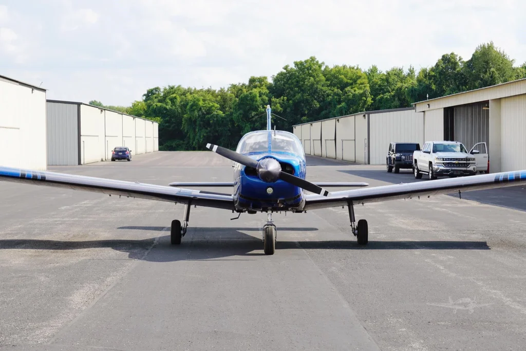 Front view of a blue single-engine propeller airplane parked on a taxiway with white hangars and green trees in the background