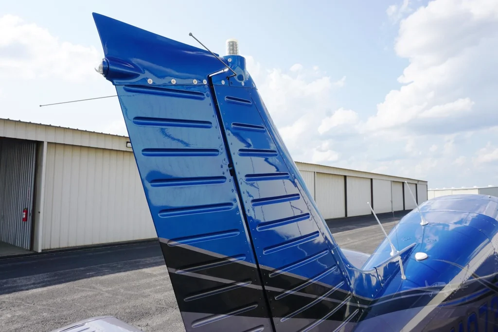 Close-up of a glossy blue airplane tail fin with rivets, set against a blue sky near a hangar.