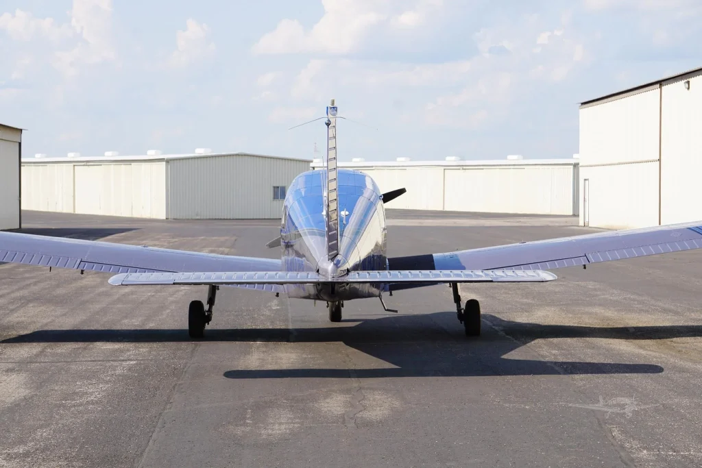Front view of a small shiny silver airplane parked on a tarmac with white hangars in the background and a blue sky with clouds above.