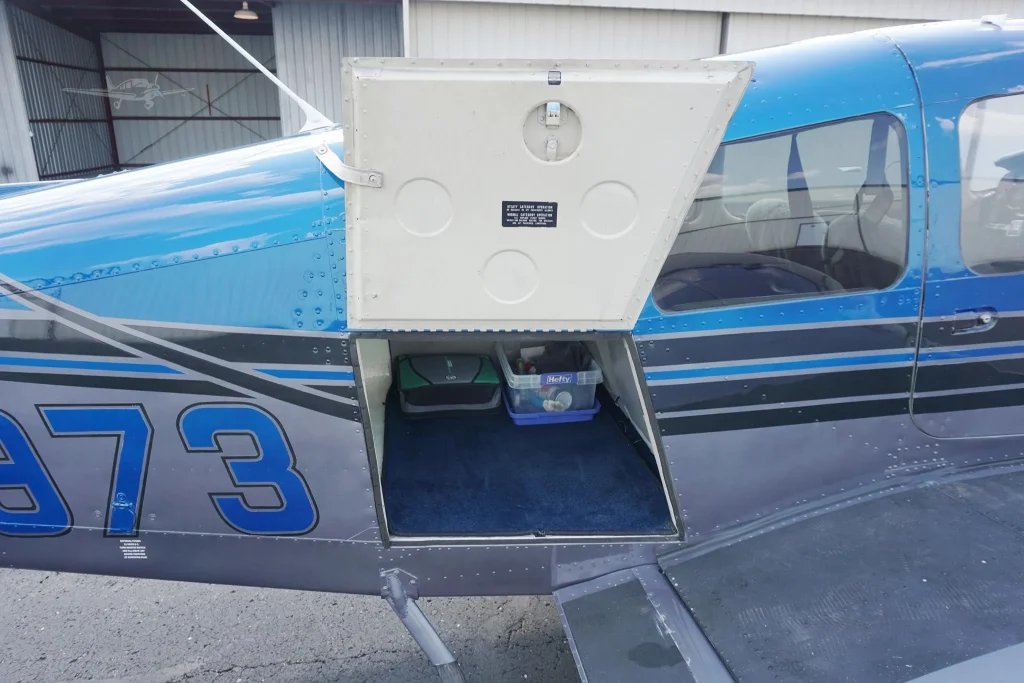 Open cargo door of a small blue airplane, revealing a plastic tote and equipment inside the baggage compartment