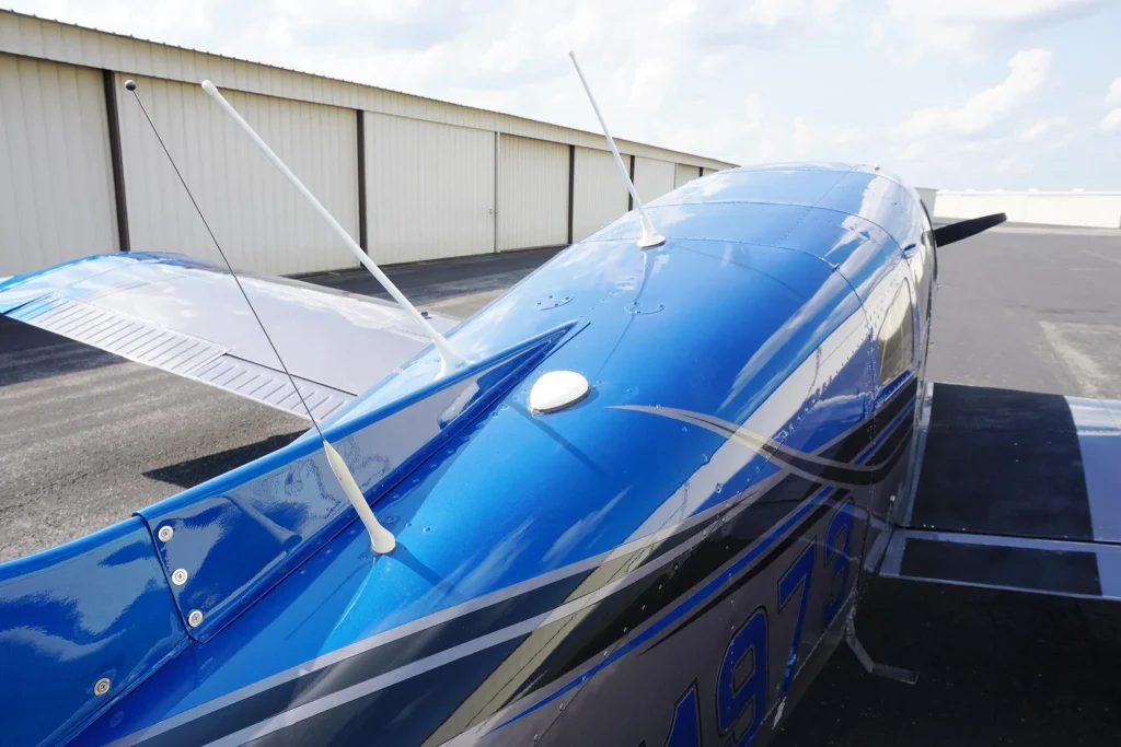 Blue single-engine airplane parked on a tarmac beside a row of hangars, antennas on fuselage visible during daytime rain-free weather