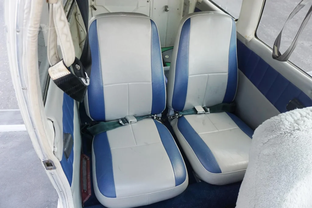 Two blue-gray airplane seats facing forward inside a small cockpit with seat belts fastened and worn upholstery.