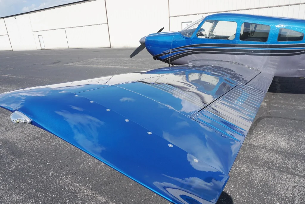Blue single-engine airplane parked on a tarmac beside a white hangar, seen from the wingtip area with a visible propeller.