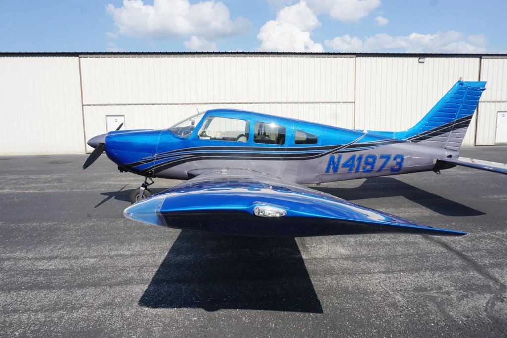 Blue single-engine propeller plane parked on a tarmac in front of a beige hangar, with a clear blue sky above and registration N41973 visible on the fuselage.