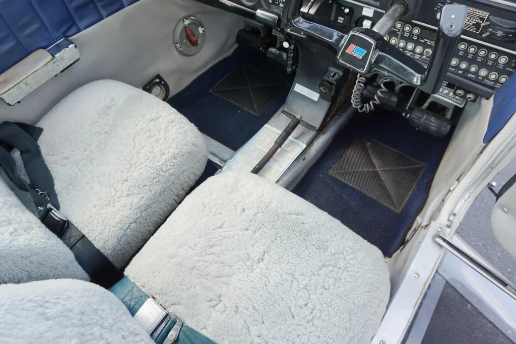 Interior of a small airplane cockpit with plush beige seats, blue carpet, and a central control yoke with throttle levers nearby.