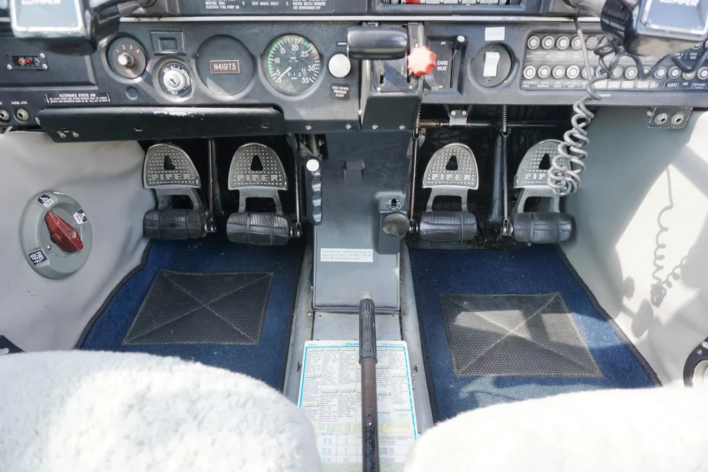 Cockpit view showing three toe-operated rudder pedals labeled 'PIPER' beneath the instrument panel, with a center console and control throttle in the middle.