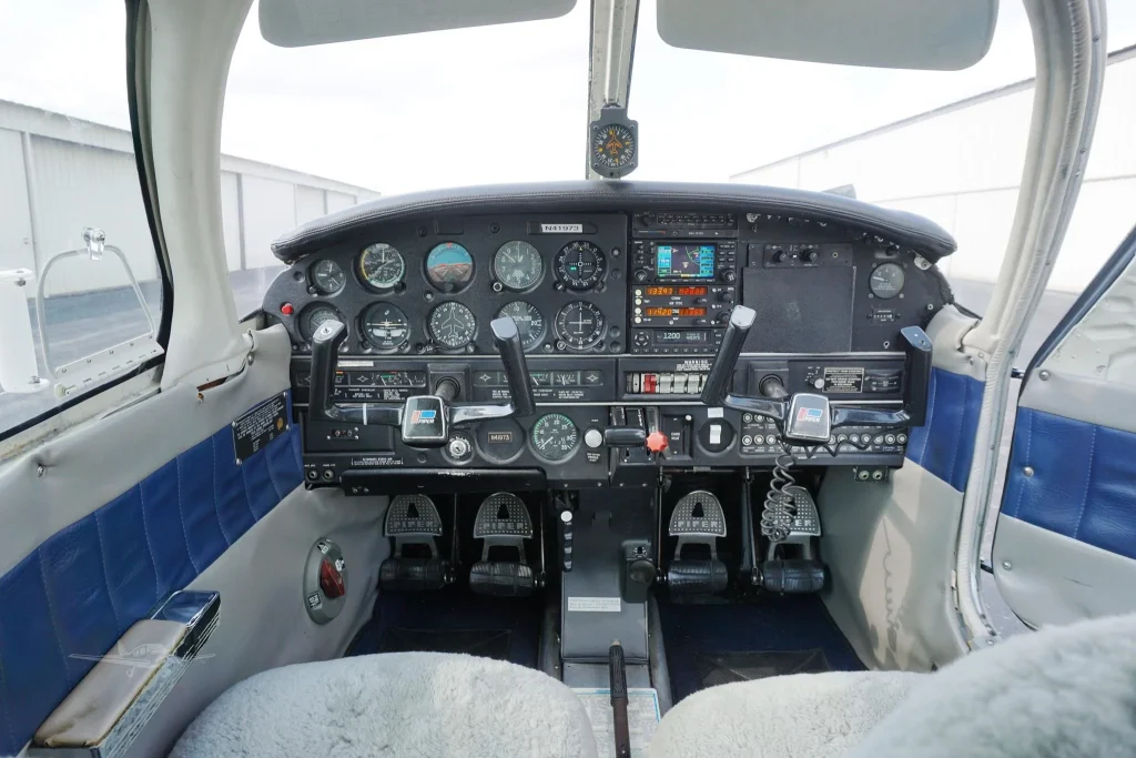 Front cockpit view of a small propeller airplane, showing numerous gauges, radios, and two control yokes on a dark panel.