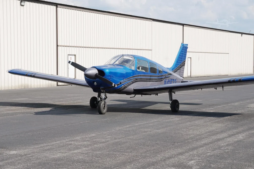 Blue single-engine propeller airplane parked on the tarmac next to a white corrugated hangar, cockpit windows visible.