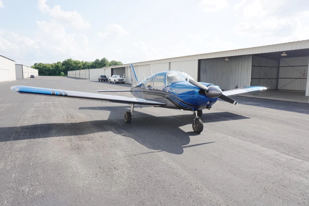 Blue single-engine propeller airplane parked on an airport tarmac beside open hangar doors under a sunny sky.